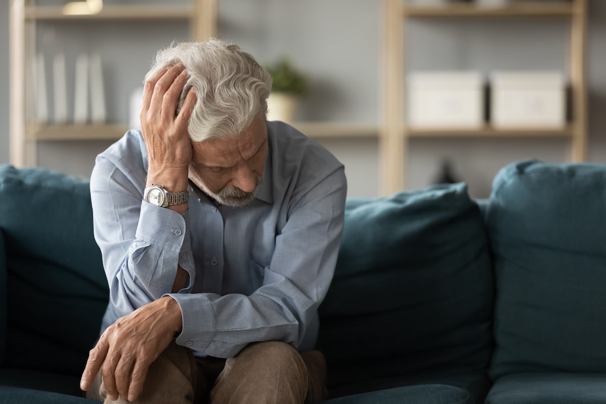 Older adult sitting on a couch touching his head.